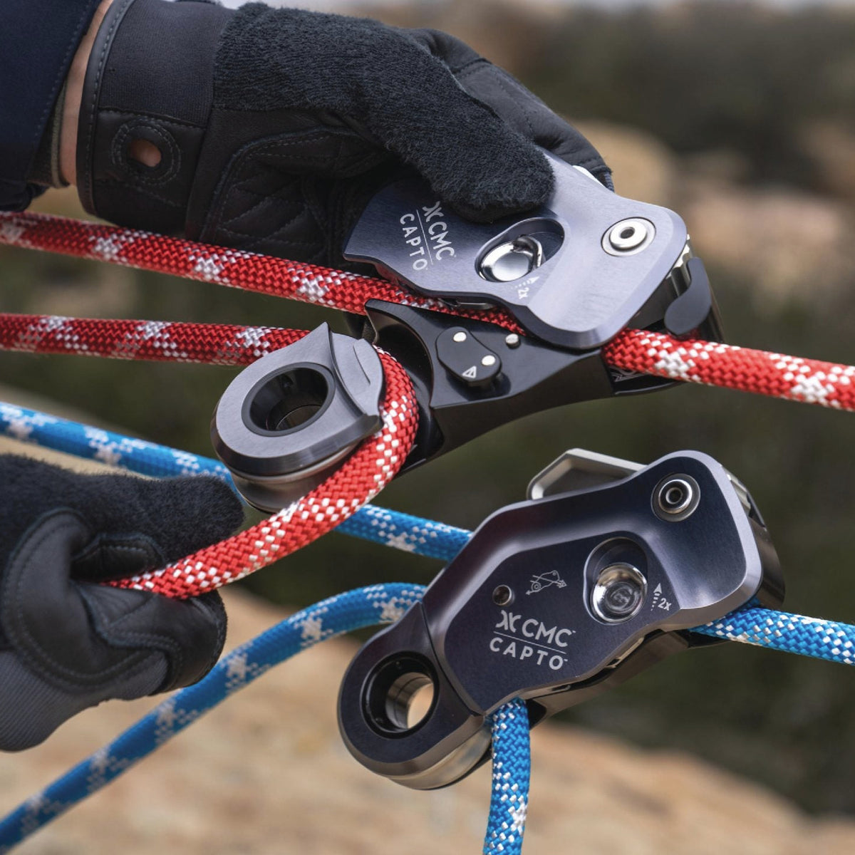 Close-up of two gloved hands handling climbing ropes and black pulleys outdoors. The ropes are red-and-white and blue-and-white, and the pulleys show the brand name ZONE CARRO.