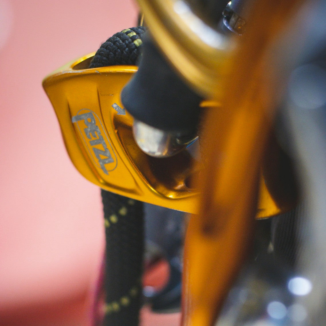 Close-up of a climbing gear component, featuring a bright orange anodized metal piece with the brand name 'petzl' printed in blue, alongside a black rope and dark gray fabric.