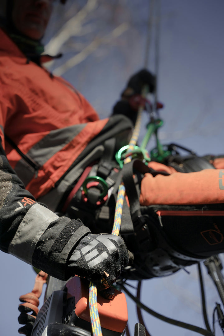 A close-up action shot of a person in an orange and gray climbing jacket wearing a harness and gloves while handling climbing ropes and gear against a blue sky.