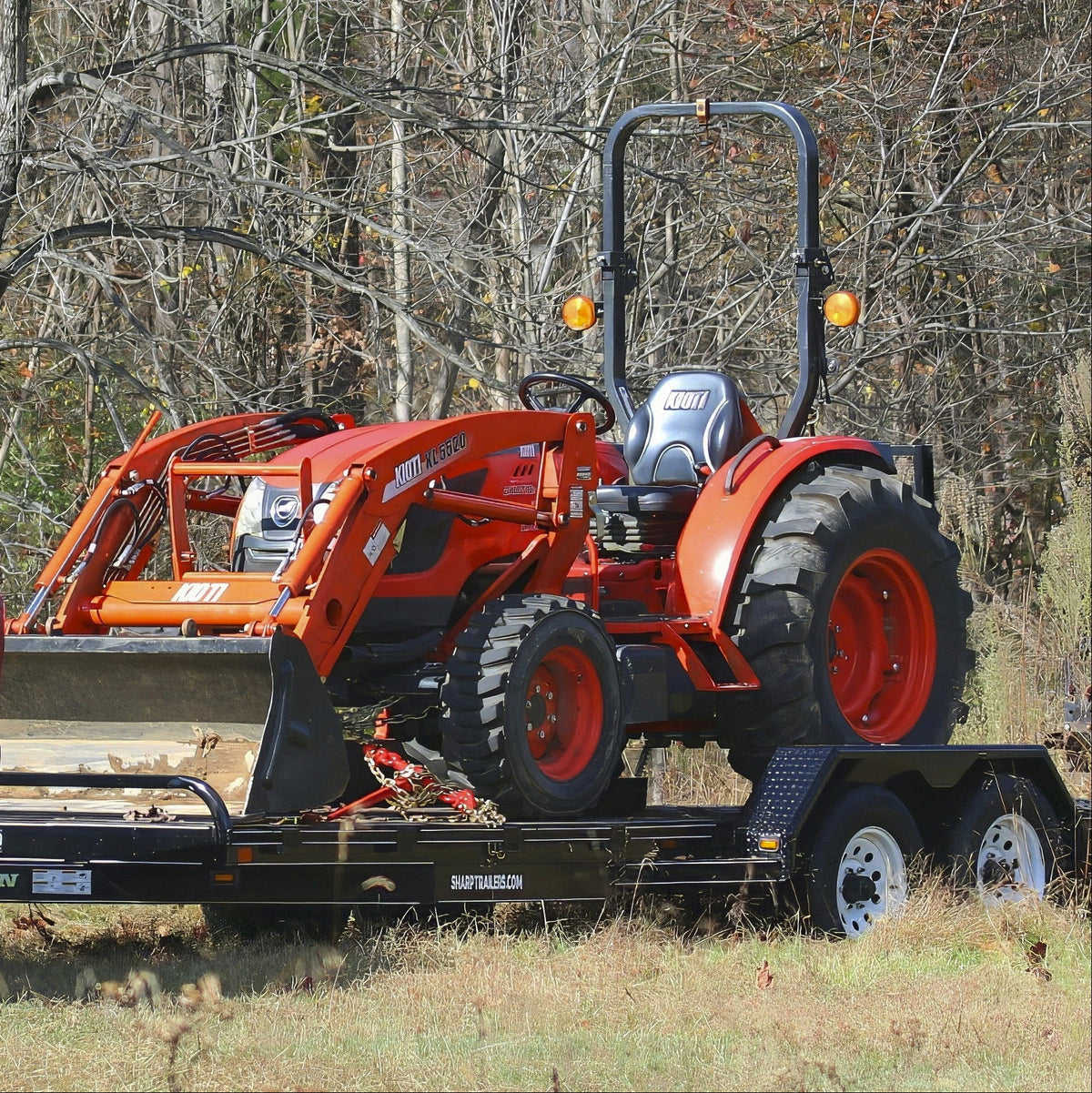 An orange Kioti tractor with a front loader attachment parked on a black flatbed trailer outdoors, with bare trees and grass in the background.