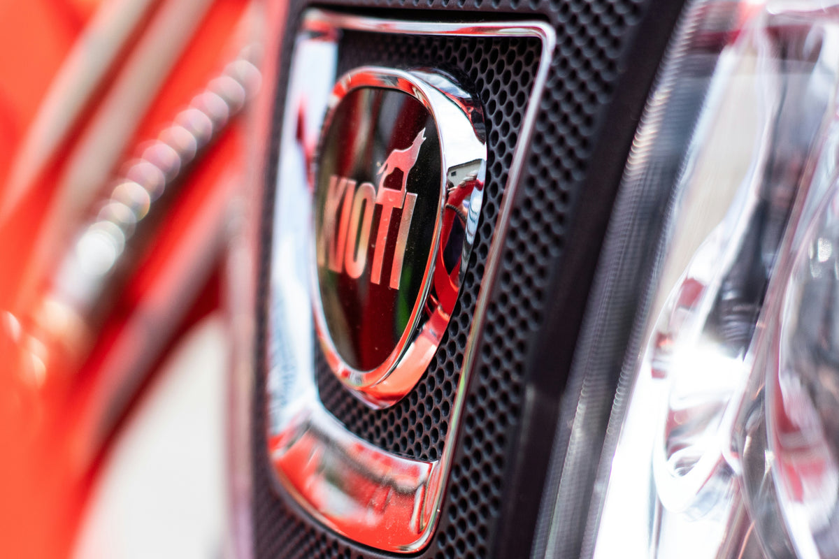 Close-up of a shiny chrome Kiwi logo badge on a black honeycomb grille, with a blurred red vehicle body in the background.