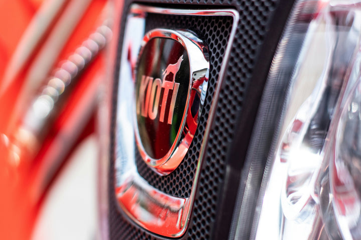 Close-up of a shiny chrome Kiwi logo badge on a black honeycomb grille, with a blurred red vehicle body in the background.